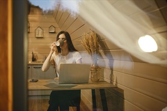 View through the window at a woman sitting at a table and drinking tea in wooden house in the