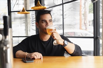 A young Latin man in a black shirt tastes a lager beer at a wooden bar table in a modern bar with