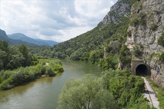 Natural riverbank at the foot of an impressive rock face under a cloudy sky, railway tunnel, Nestos