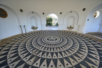 A detailed mosaic on the floor of a white circular building with open arches to the outside,