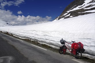 A bicycle with red panniers stands next to a snow-covered mountain road under a clear sky, Fjell in