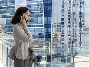 A woman is standing on a balcony, holding a paper cup of coffee and talking on her cell phone