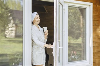 Smiling woman in bathrobe and cup of coffee in her opens the door onto terrace of wooden cabin