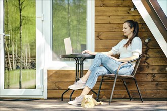 Relaxed woman sitting with laptop on terrace of log cabin in a sunny day