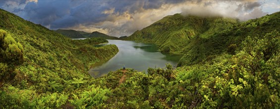 Panoramic view of a lake surrounded by green forested mountains under a dramatic sky, crater lake