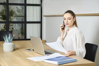 Businesswoman sitting at a desk in office, holding a document and speaking on the phone with