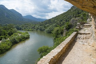 Hiking trail under a rock overhang overlooking a calm river and green treetops, River Nestos or