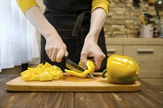 Unrecognizable man in apron slicing bell peppers on wooden cutting board in the kitchen