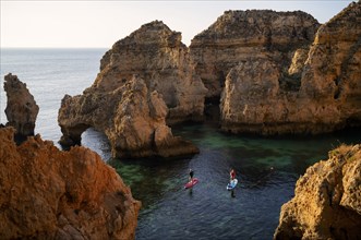 Stand Up Paddlers on their boards, colourful cliffs and sunrise, rocks in the sea, Ponta da