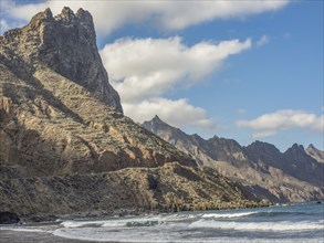 Mighty rock formations on the coast reaching down to the sea, under a partly cloudy blue sky,