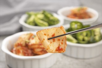 A food photo of a pair of chopsticks holding a piece of kimchi above bowls of kimchi and sliced