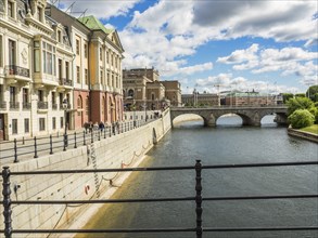 View of a waterfront promenade with historic buildings along a river, a bridge and a partly cloudy