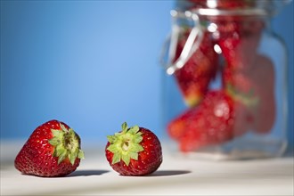 Close up of two strawberries on a table with blue background and a glass jar full of strawberries