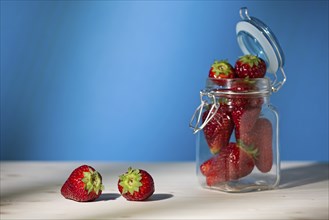 Strawberries on a table and a glass jar full of strawberries with blue background