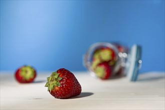 Close up of a strawberry and a glass jar full of strawberries lying down on a table with blue