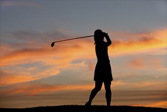 A silhouette of a woman golfer at sunset swinging the golf club