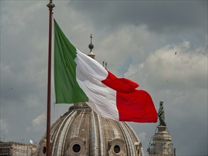 The Italian flag flies in front of a dome and a sculpture under a cloudy sky, rome, italy