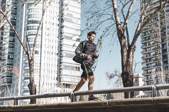 Side view of motivated handsome man jogging by green public park. Copy space