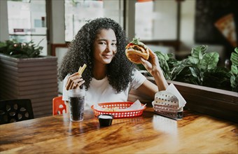 Happy girl enjoying a delicious hamburger sitting in a restaurant. Portrait of young afro woman