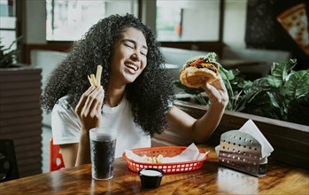 Happy girl enjoying a hamburger in a restaurant. Afro young woman enjoying a hamburger with fries