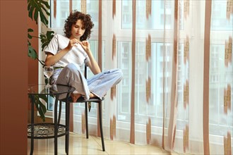 Woman examining broken fingernail sitting on chair by the window