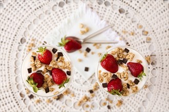 Strawberries cereals and chocolate flakes in two cups of plain yogurt seen from above