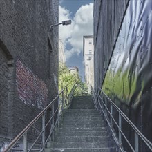 A staircase leads through a narrow alley into bright sunlight, flanked by brick walls and steel