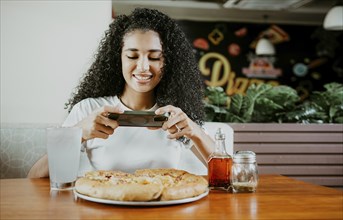 Smiling young woman using phone photographing a pizza in a restaurant. Happy girl photographing a