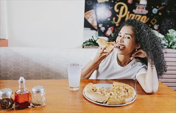 Smiling young woman enjoying a pizza in a restaurant. Happy afro hair girl eating pizza in a