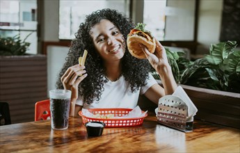 Afro young woman enjoying a hamburger with fries in a restaurant. Happy girl enjoying a hamburger