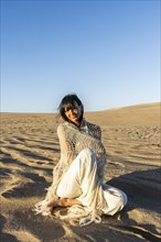 A woman dressed in white sitting on the sand at sunset