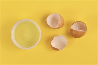 Bowl with egg white next to broken eggshells on yellow background