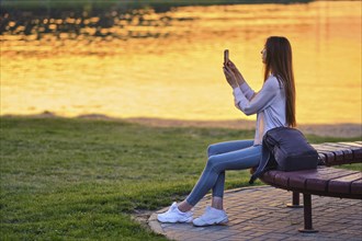 Blogger woman streaming online the beautiful sunset sitting on bench by river