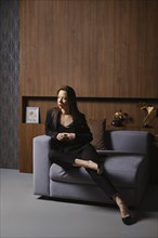 Full length portrait of an attractive woman in a black suit sitting in armchair in a modern