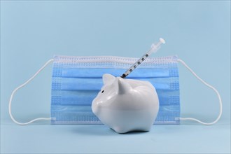 Corona virus vaccine expenses concept with white piggy bank, syringe and medical face mask on blue