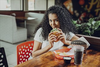 Portrait of afro girl holding a hamburger in a restaurant. Beautiful latin girl enjoying a