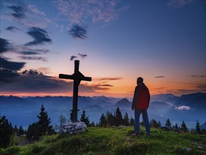 Mountaineer standing next to the summit cross of the Ahornbüchsenkopf at dawn, Roßfeld,