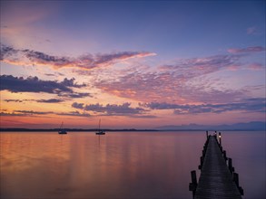 Illuminated clouds over sailing boats and jetty at Chiemsee at dawn, behind the Chiemgau Alps,