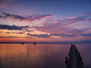 Illuminated clouds over sailing boats and jetty at Chiemsee at dawn, behind the Chiemgau Alps,