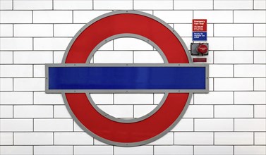 A well-known round tube sign in red and blue on a white tiled wall of a railway station, London,