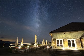 A mountain hut under a clear starry sky with the Milky Way visible and mountains in the background,