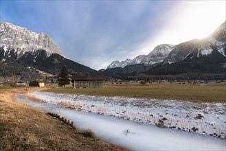 Frozen river on a meadow with dried grass, wooden cabins, a village, and the snow-capped Alps