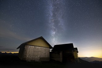 A mountain hut under a clear starry sky with the Milky Way visible and mountains in the background,