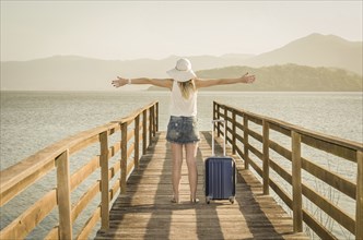 Great vacation concept. Young woman with open arms and suitcase waiting for the boat on the dock