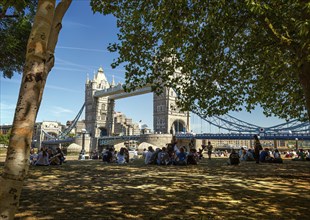 The historic Tower Bridge, in front of it a shady park where people sit in the shade of trees,