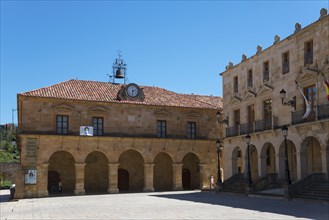 A quiet square with historic buildings, including a clock tower and arcades, under a clear blue