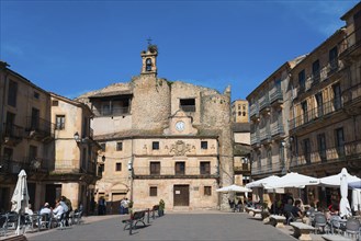 Historic square with stone walls, cafés and parasols, bustling with people and dominated by a clock