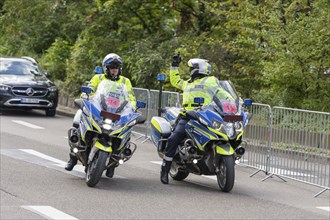 Two policemen on motorbikes in uniform on a street surrounded by security barriers and trees,