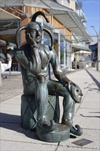 Bronze statue of a pensive jester sitting on a cube and holding a mask next to him, carnival jester