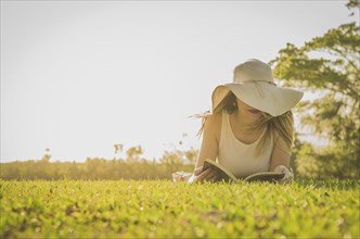 Pretty woman reading book lying on the lawn, seen from above with summer hat